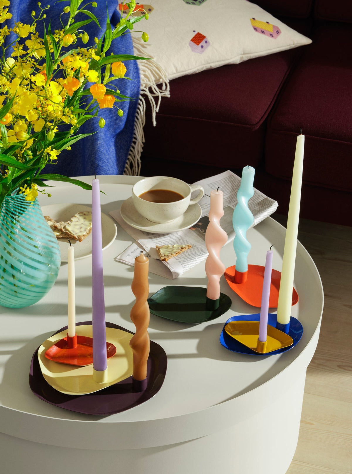 Thin taper candles in coloured holders on a round table with yellow flowers