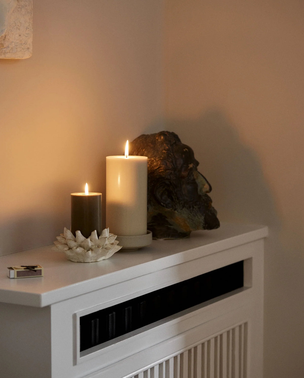 row of lit Nordal pillar candles on a white mantelpiece shelf, two tall cream candles, a shorter cream candle, two dark olive green candles of varying heights, another tall cream candle and a small dark candle, with framed artwork on the wall behind