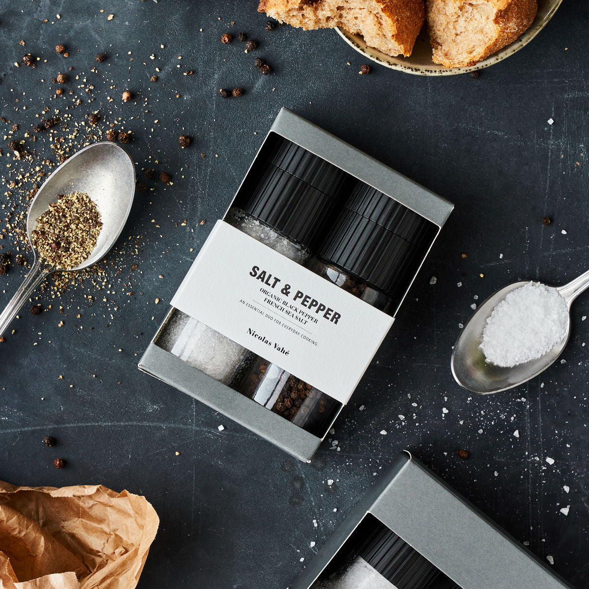 Salt and pepper shakers in a box on a dark surface with spoons and bread.