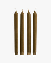 row of lit candles on a white mantelpiece shelf: two ceramic fish-shaped candlesticks holding olive green taper candles, a cluster of cream and dark olive green pillar candles of varying heights, and a brass candlestick holding an olive green taper candle, with framed artwork on the wall behind