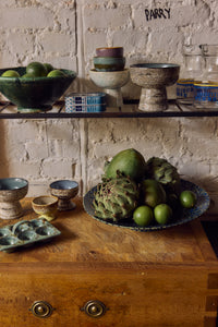 70s Ceramics tapas bowl on base in Brew displayed on an open shelf with artichokes, olives and other kitchen items