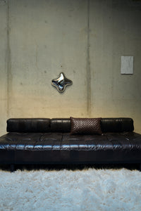 Chocolate brown woven cushion styled on a dark leather sofa against a concrete wall
