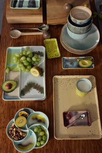 The HKLIVING Tide cake tray as part of an overhead flat lay tablescape, styled with grapes, avocado, nuts and other 70s ceramics trays and bowls on a wooden table