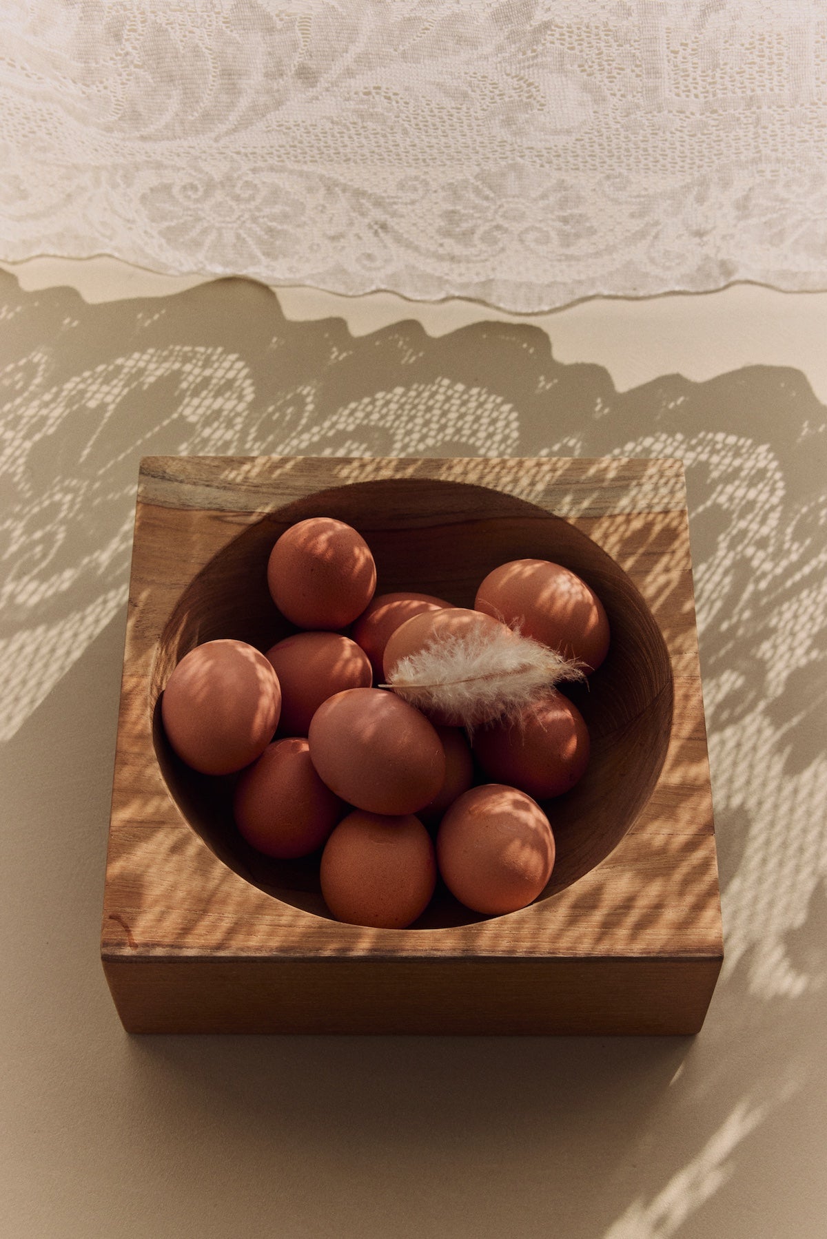 HKLIVING teak cube bowl filled with brown eggs and a feather, styled beneath a lace curtain casting dappled shadow