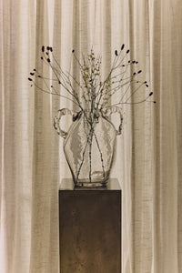 Clear glass amphora hkliving vase with dried branches styled on a metal plinth against linen curtains