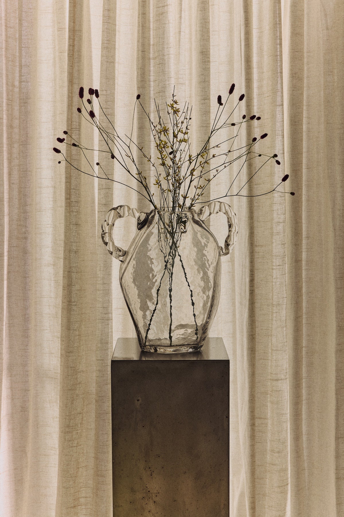 Clear glass amphora hkliving vase with dried branches styled on a metal plinth against linen curtains