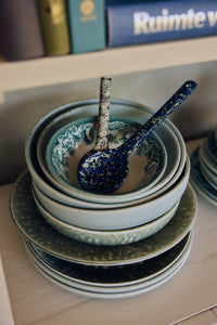 Close-up of 70s ceramics serving spoons in Blues resting in a patterned bowl atop a stack of blue-toned dinnerware