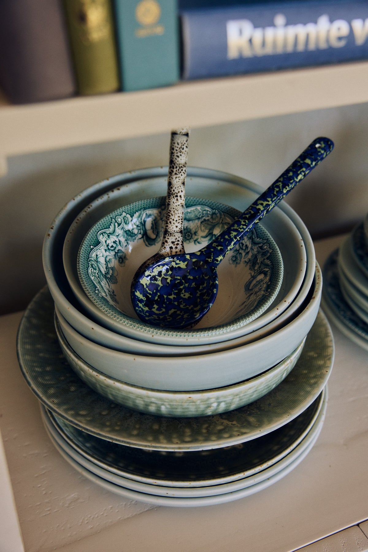 Close-up of 70s ceramics serving spoons in Blues resting in a patterned bowl atop a stack of blue-toned dinnerware