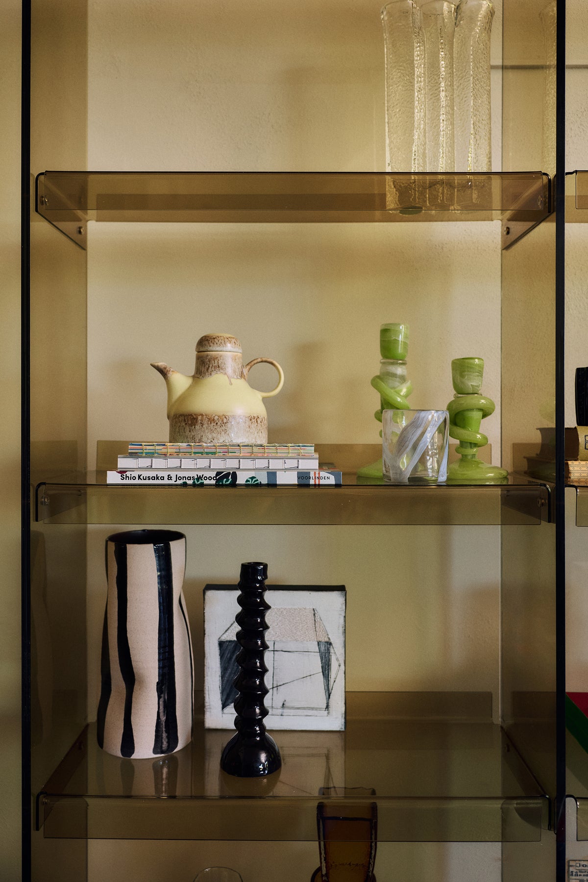 The Mono Vase, Sand/Black by HKLIVING styled on open glass shelving with a black ribbed candlestick, next to a yellow ceramic pitcher and green glass candlesticks above