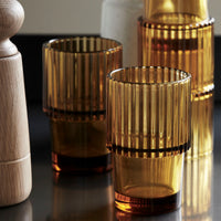 Set of Rills amber drinking glasses styled with a wooden pepper mill on a dark kitchen surface, House Doctor fluted tumblers ready for everyday dining