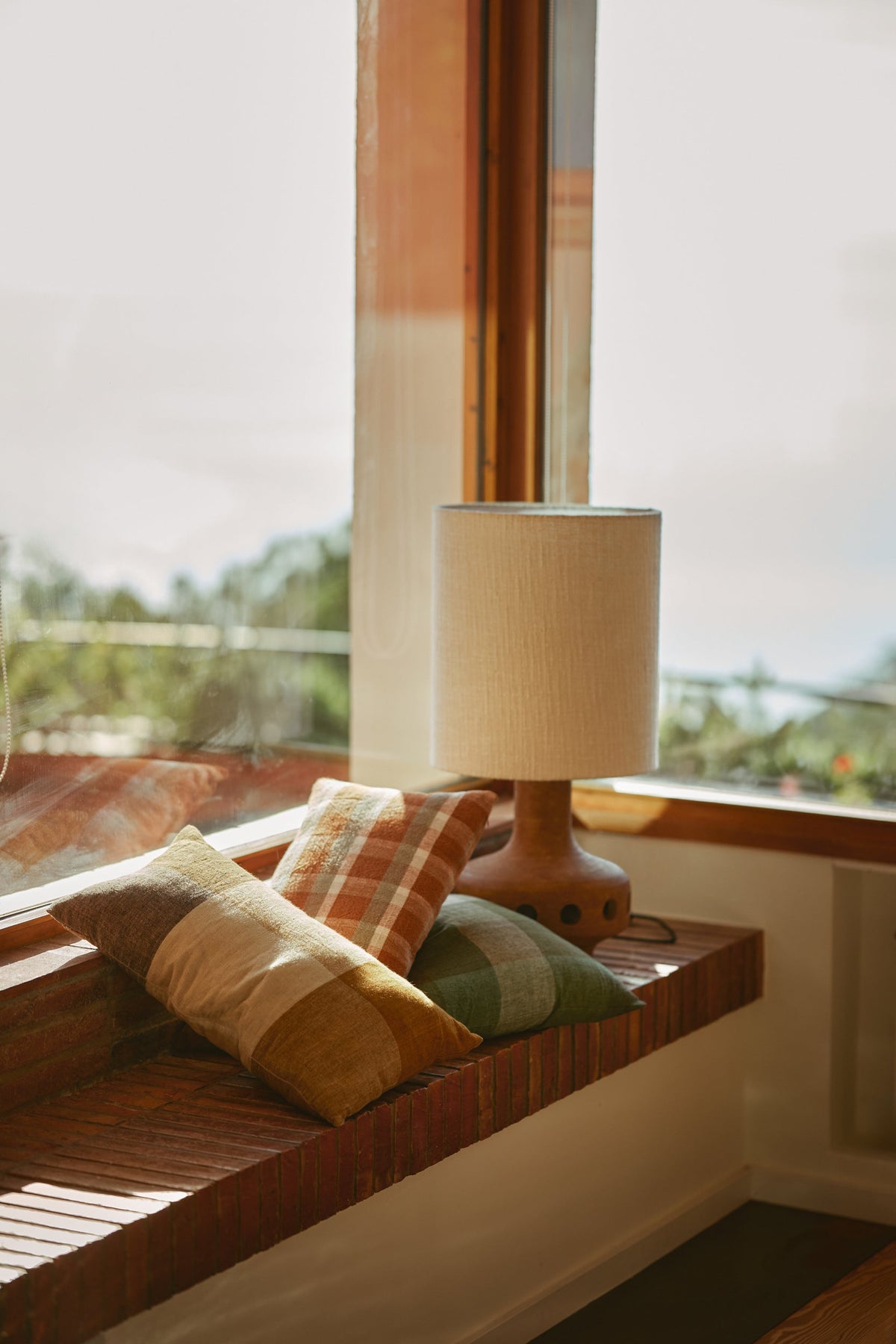 Wooden bench with cushions and a lamp by a window with a view of greenery