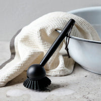 Meraki black bamboo dish brush styled beside an enamel basin and waffle linen tea towel on a stone counter, a calm Scandinavian washing-up moment