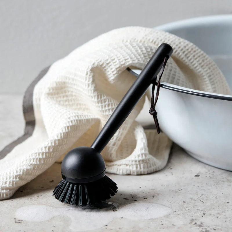 Meraki black bamboo dish brush styled beside an enamel basin and waffle linen tea towel on a stone counter, a calm Scandinavian washing-up moment