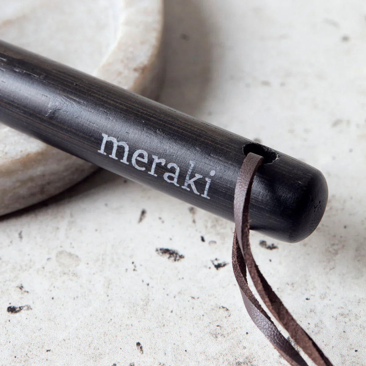 Close-up of the Meraki logo engraved on the bamboo handle of the black dish brush, with the dark leather hanging strap