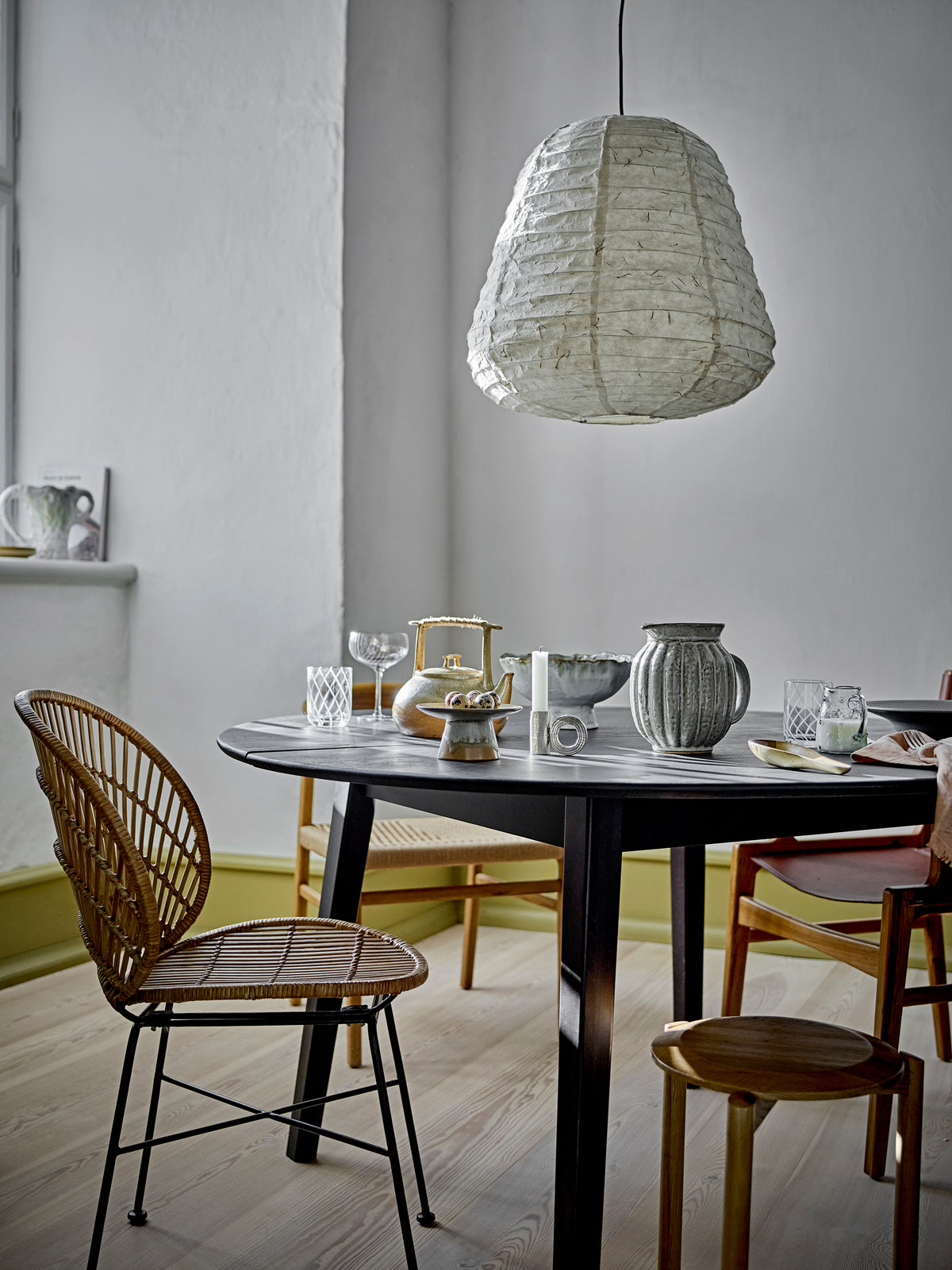 Round dining table set with glassware and stoneware beside a rattan chair, under a white paper pendant light