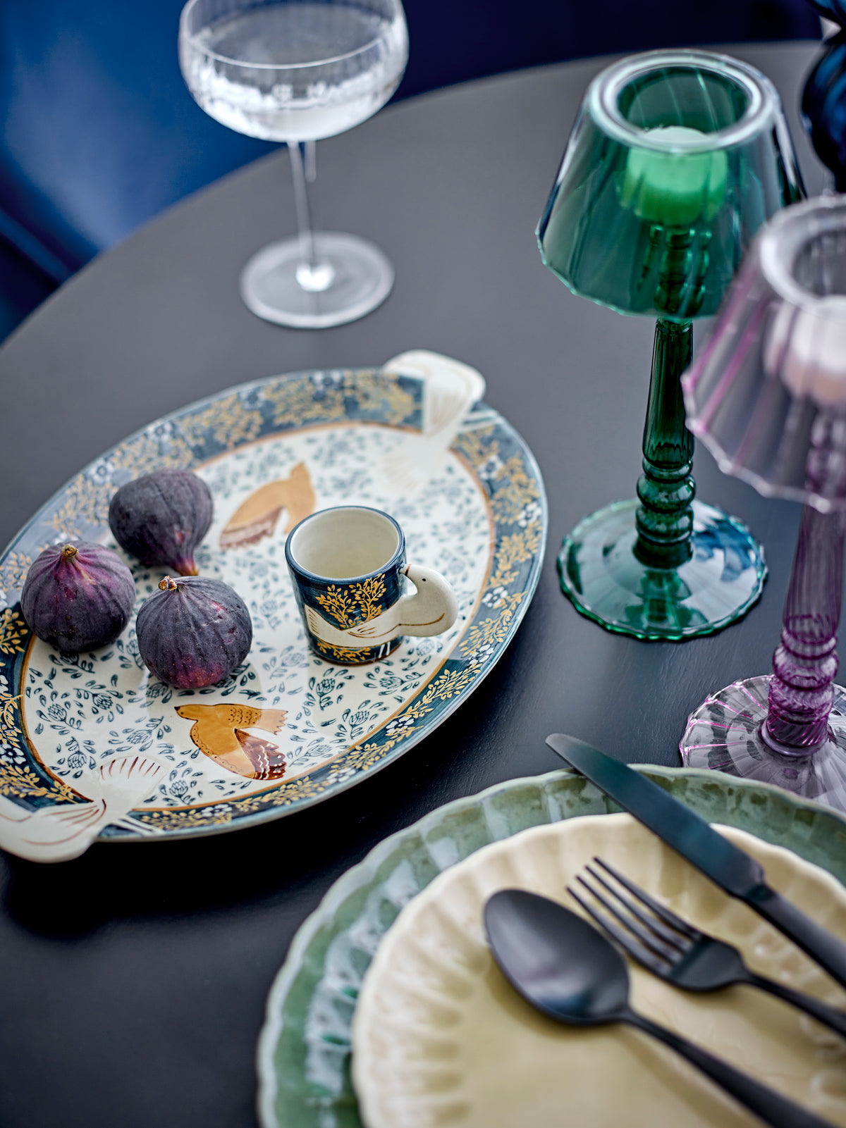 Dark round table set with a patterned oval platter, figs, a small espresso cup, a green glass candle holder and a coupe glass