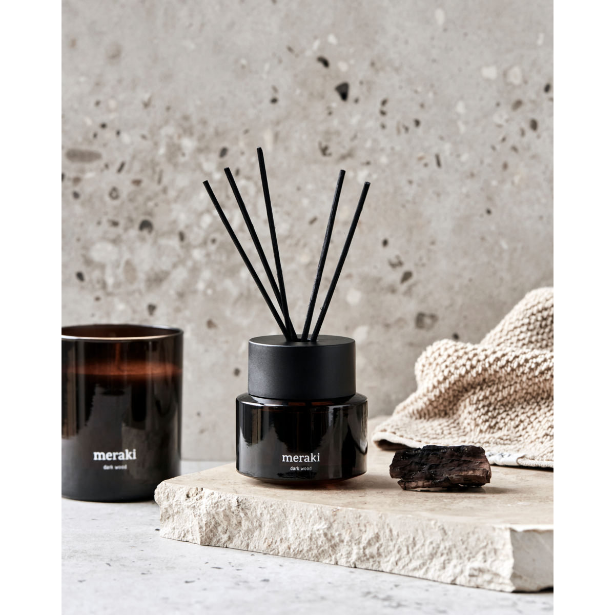 Meraki Dark Wood reed diffuser beside a Dark Wood candle on a stone ledge with a linen cloth