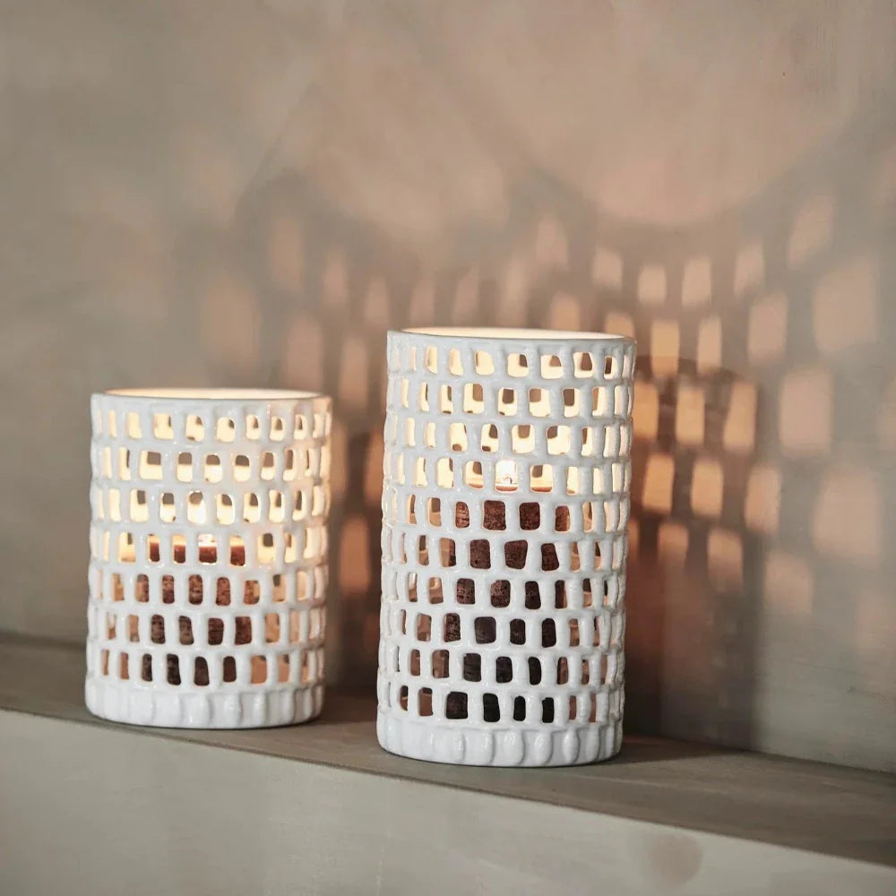 2 white cearmic candle holders on a mantle piece with pillar candles glowing inside seen through the hand cut holes all around the pieces