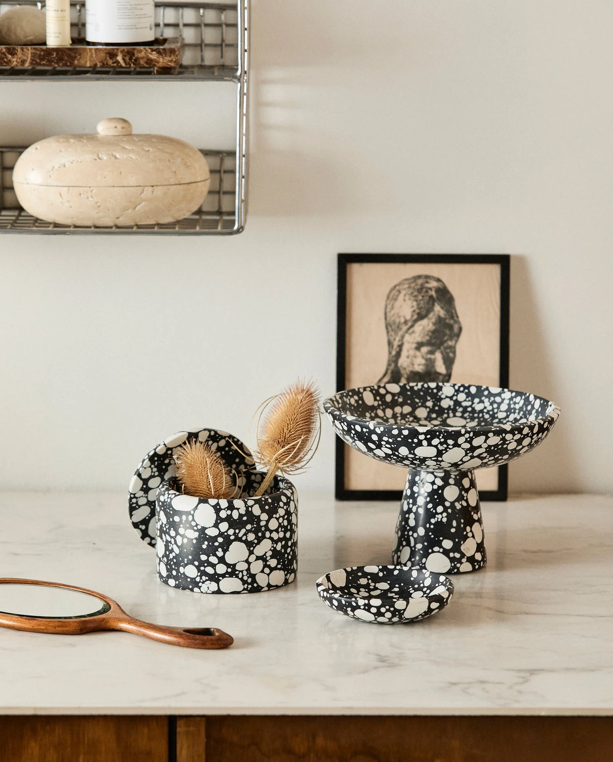 black and white terrazzo bowls and a small dish styled on a white marble counter with a travertine trinket box, a wooden hand mirror and a framed print in the background
