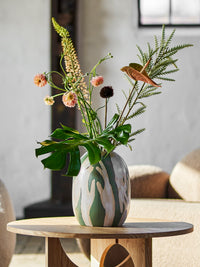 Adalena green vase styled on a wooden side table with foliage and pink flowers