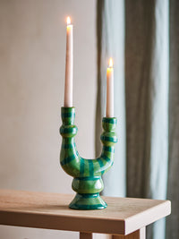 Salandy green striped candle holder with two lit taper candles on a pale wooden console table in front of a grey curtain