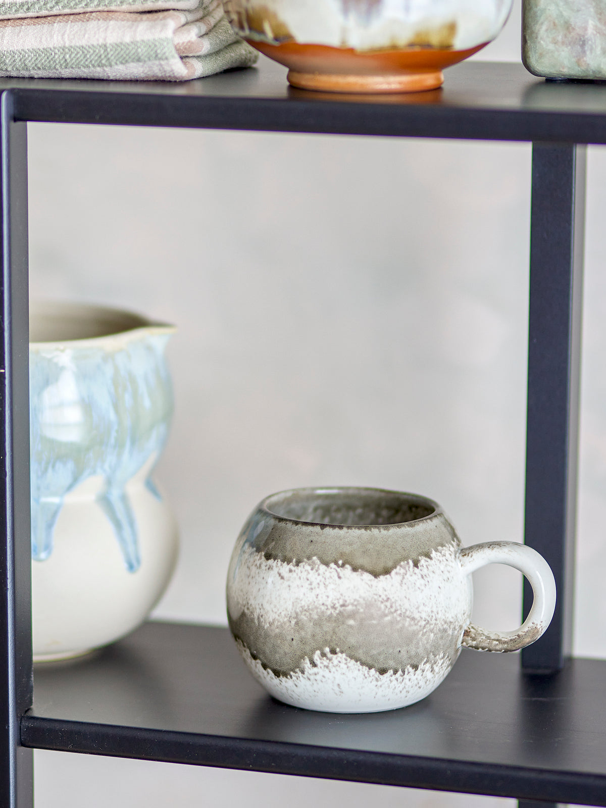Paula grey and cream reactive glaze mug on a black shelf beside a pale blue and white jug and folded linen