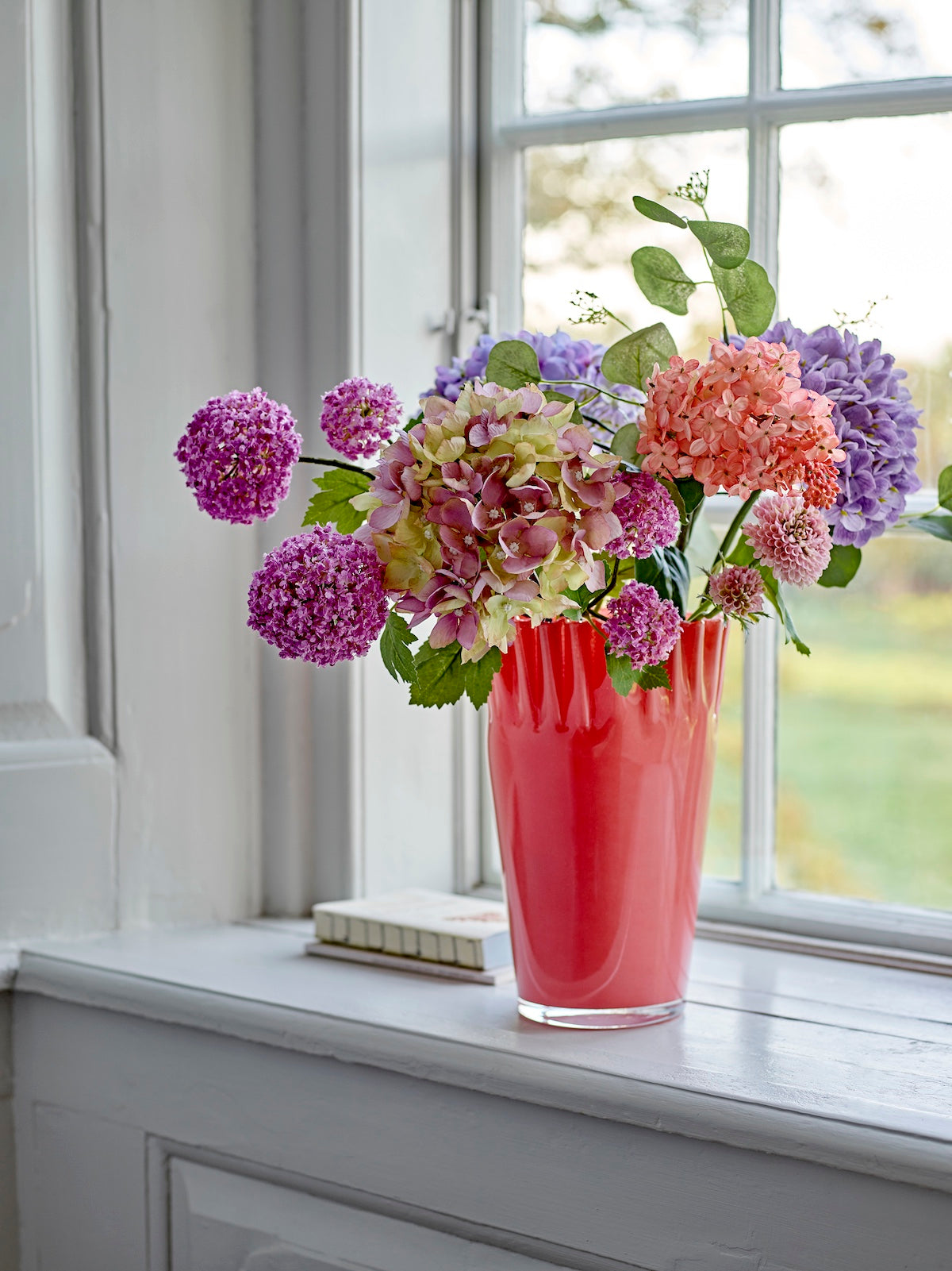 Janine coral red glass vase on a windowsill filled with purple and pink flowers