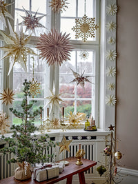 Cedar tree with a wrapped base beside gift parcels on a wooden bench in a bay window hung with paper stars and snowflakes
