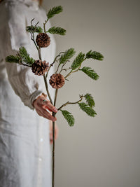 Hand holding the Pine Stem with three pine cones against a soft grey wall, beside a white linen shirt