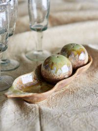 Kayo green stoneware salt and pepper shakers resting in their pea pod tray on natural linen, with clear glassware behind