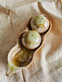 Overhead view of the Kayo salt and pepper shakers in the pea pod tray on crumpled natural linen, showing one shaker with a single hole and one with three