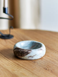 Indah grey marble bowl on a light oak table beside a black candle holder and blurred cream curtain