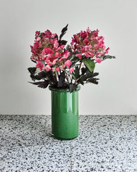 House Doctor Buck vibrant green glass vase styled on a terrazzo surface with pink hydrangeas and dark foliage, a bold confident colour pairing for modern interiors