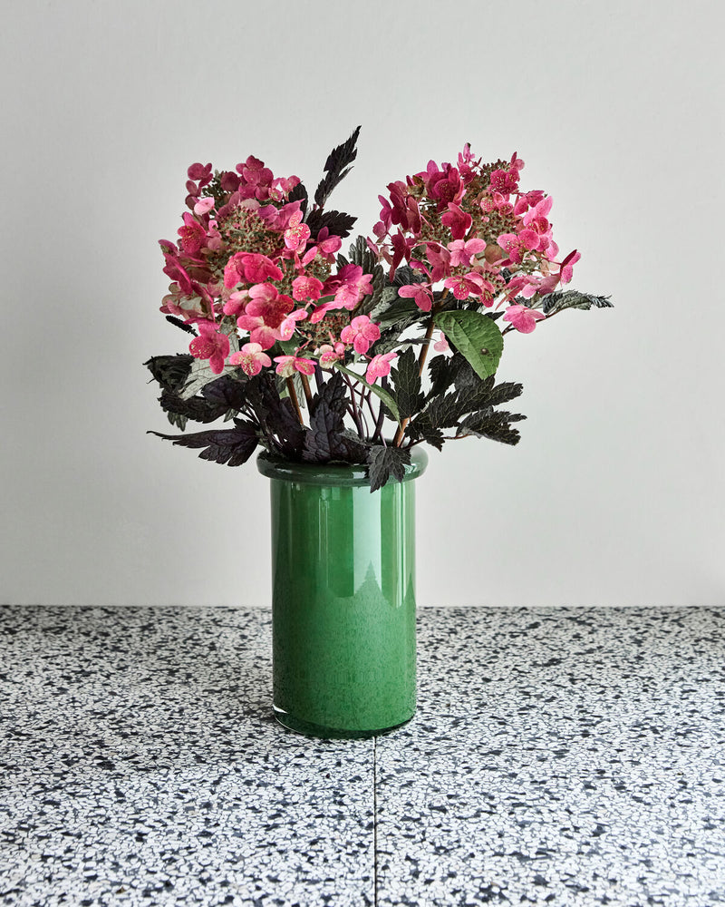 House Doctor Buck vibrant green glass vase styled on a terrazzo surface with pink hydrangeas and dark foliage, a bold confident colour pairing for modern interiors