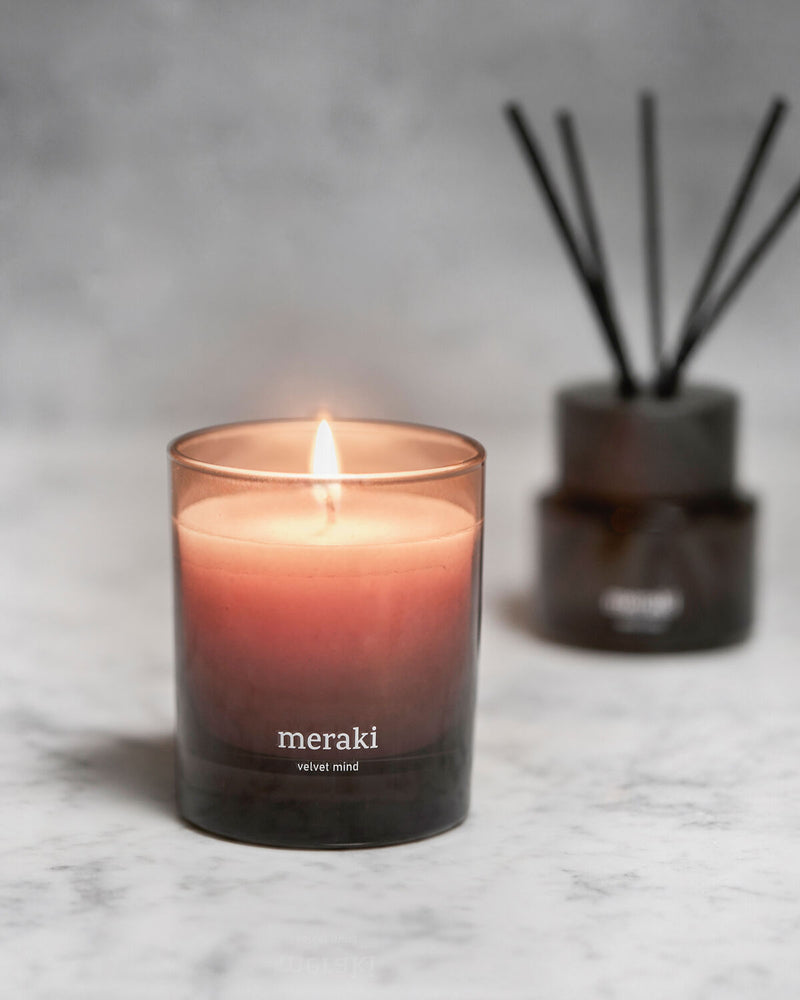 Lit Meraki Velvet Mind candle glowing warm amber through its smoked glass jar, styled on a marble surface next to a reed diffuser