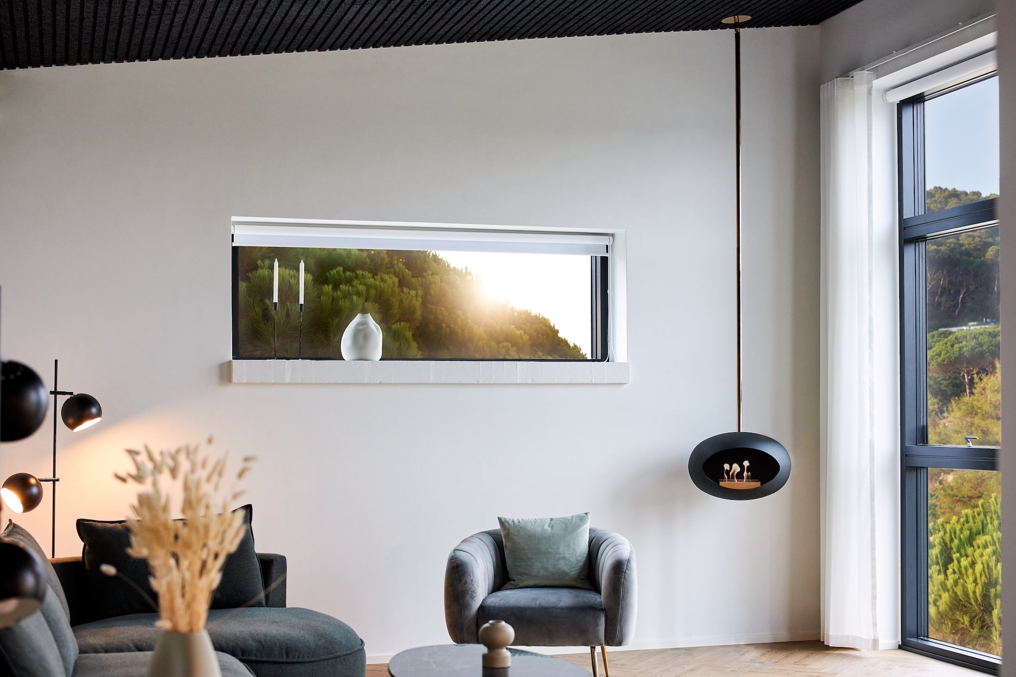 Modern living room with a large window, gray sofa, and armchair warmed by a Le Feu real flame fireplace.