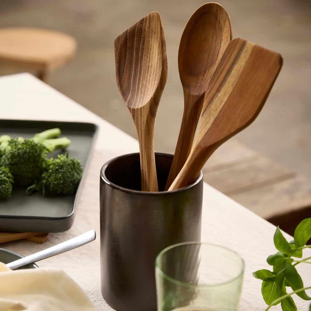 Aida wooden utensils in a black container on a kitchen counter with vegetables and a glass.