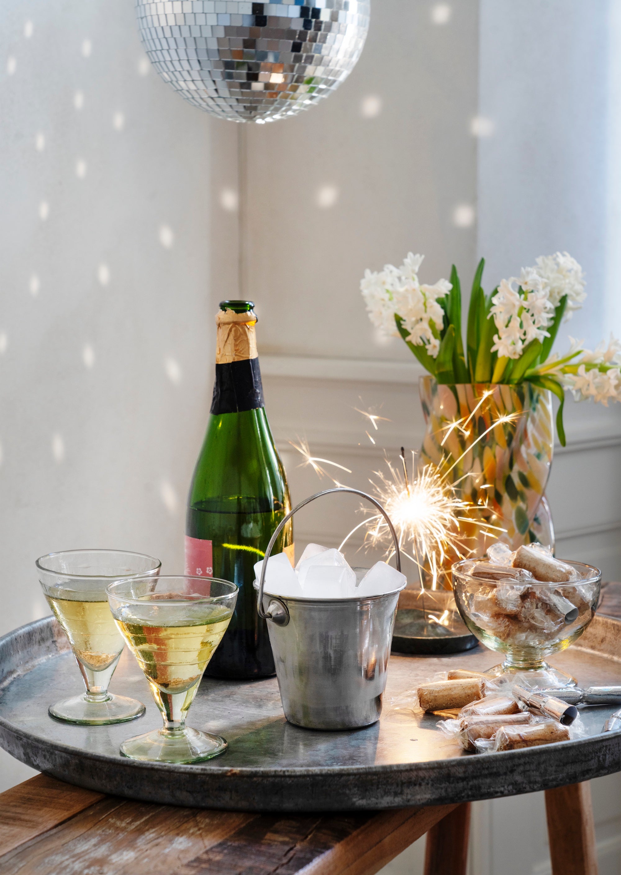 Bottle of champagne, glasses with champagne, and decorative items on a table with a disco ball in the background.