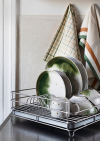 Dish rack with green and white ceramic plates and bowls, with kitchen towels hanging in the background.
