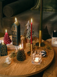 Lit dark green Casa house candles with a red tree candle, pine cone candle and brass candlesticks on a wooden coffee table