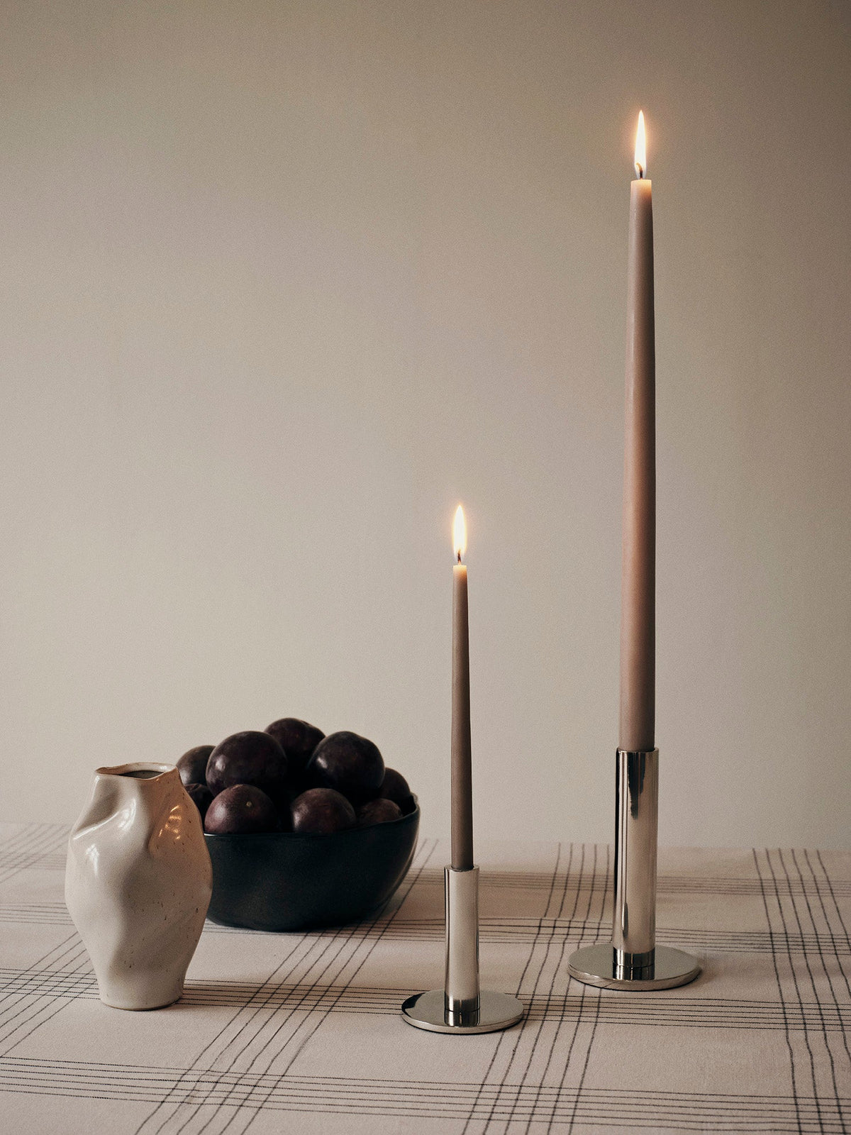Small and large silver Maro candle holders with taupe tapers beside a ceramic jug and bowl
