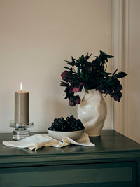 Lit pillar candle in a Petra glass holder on a dark sideboard with a white vase of flowers
