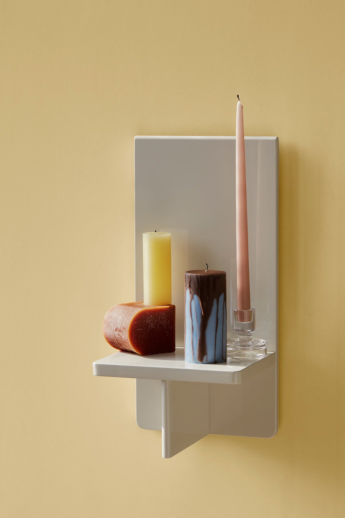 A smooth taper candle and pillar candles on a white wall shelf against a yellow wall