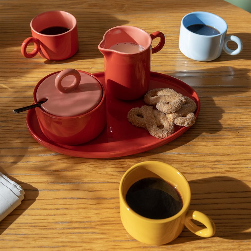 Red Chiquito milk jug, sugar pot and tray with yellow, red and blue mugs on a sunny wooden table