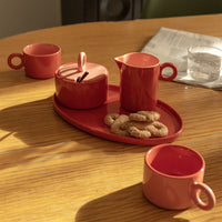 Red Chiquito tray with sugar pot, milk jug, biscuits and mugs on a sunny wooden table