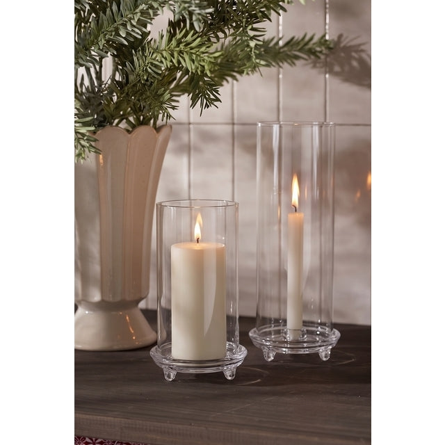 Lily glass hurricanes with lit ivory pillar and taper candles on a dark sideboard with pine sprigs