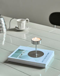 House Doctor Anit silver tealight holder with a lit tealight on a book beside a coffee cup