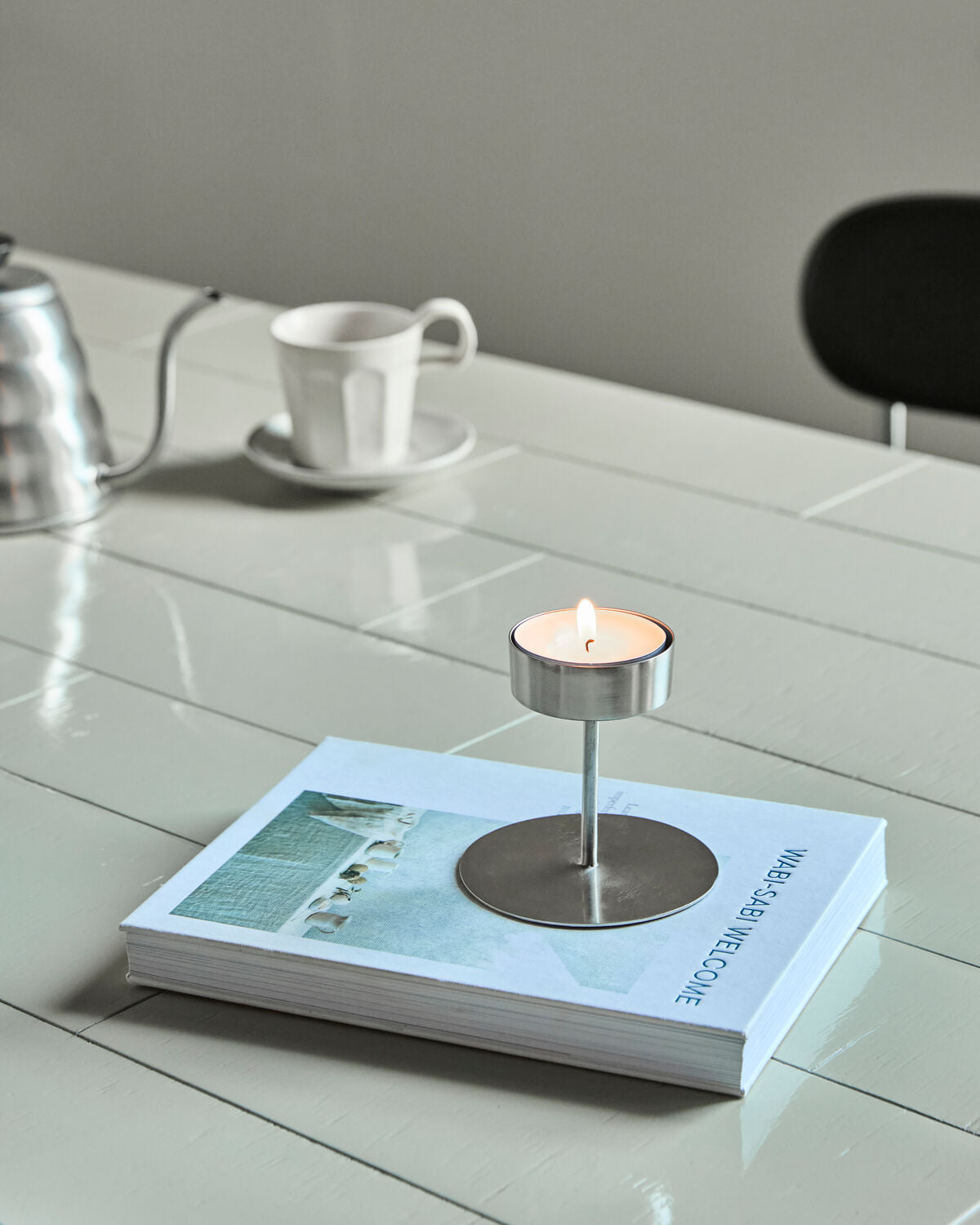 House Doctor Anit silver tealight holder with a lit tealight on a book beside a coffee cup