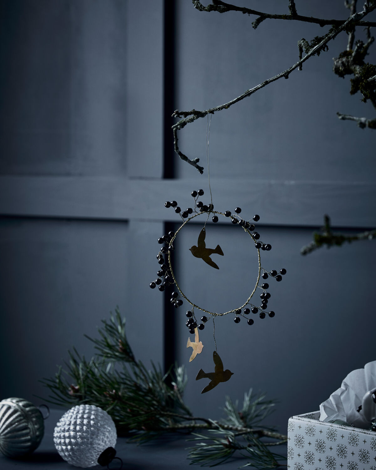 House Doctor black bead and brass bird wreath decorations hanging on bare branches against a dark blue wall