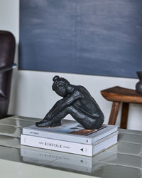 House Doctor Forms black seated figure sculpture on books on a glass table in a dark living room